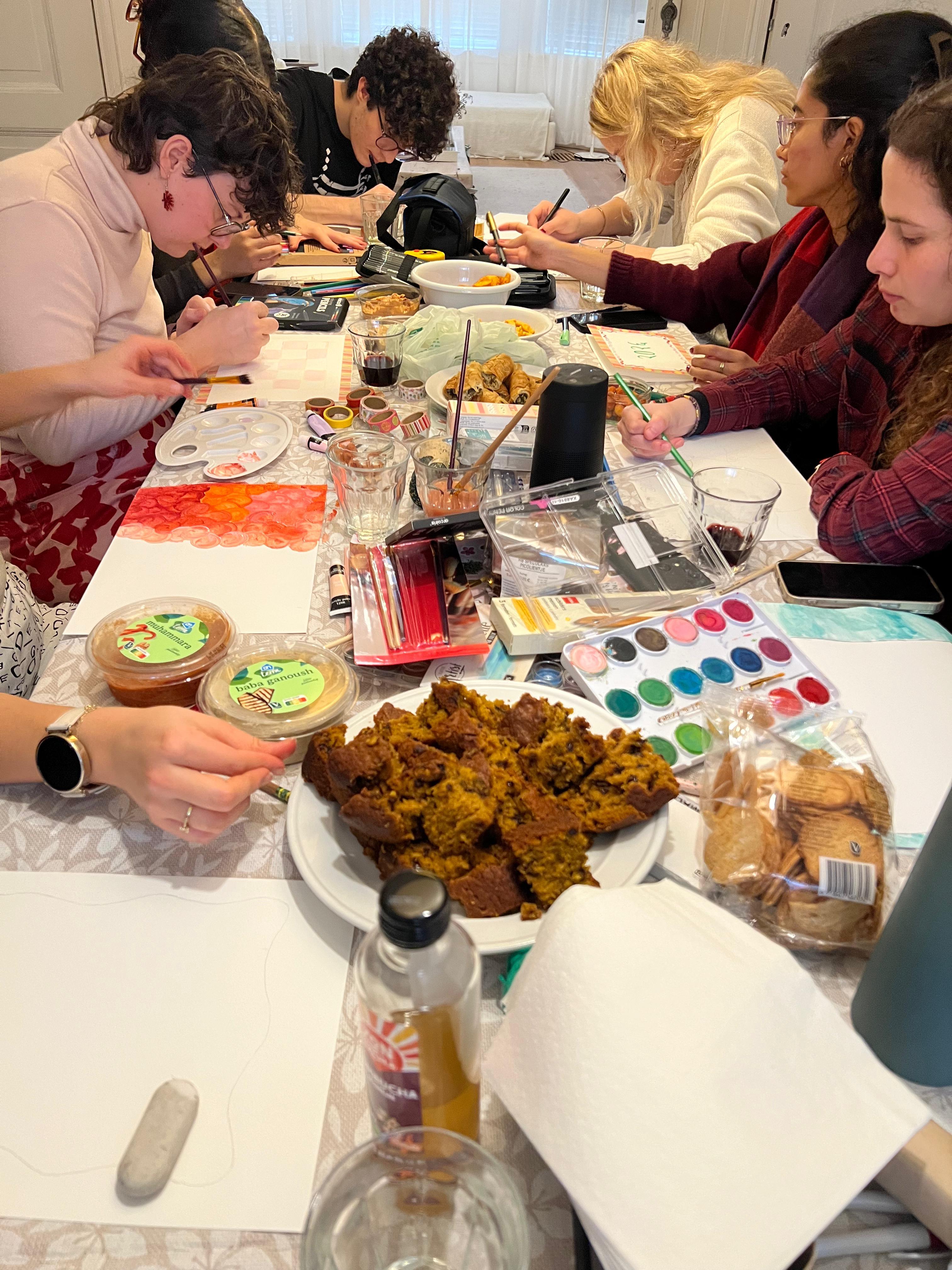Students sitting at a long table with painting articles and food